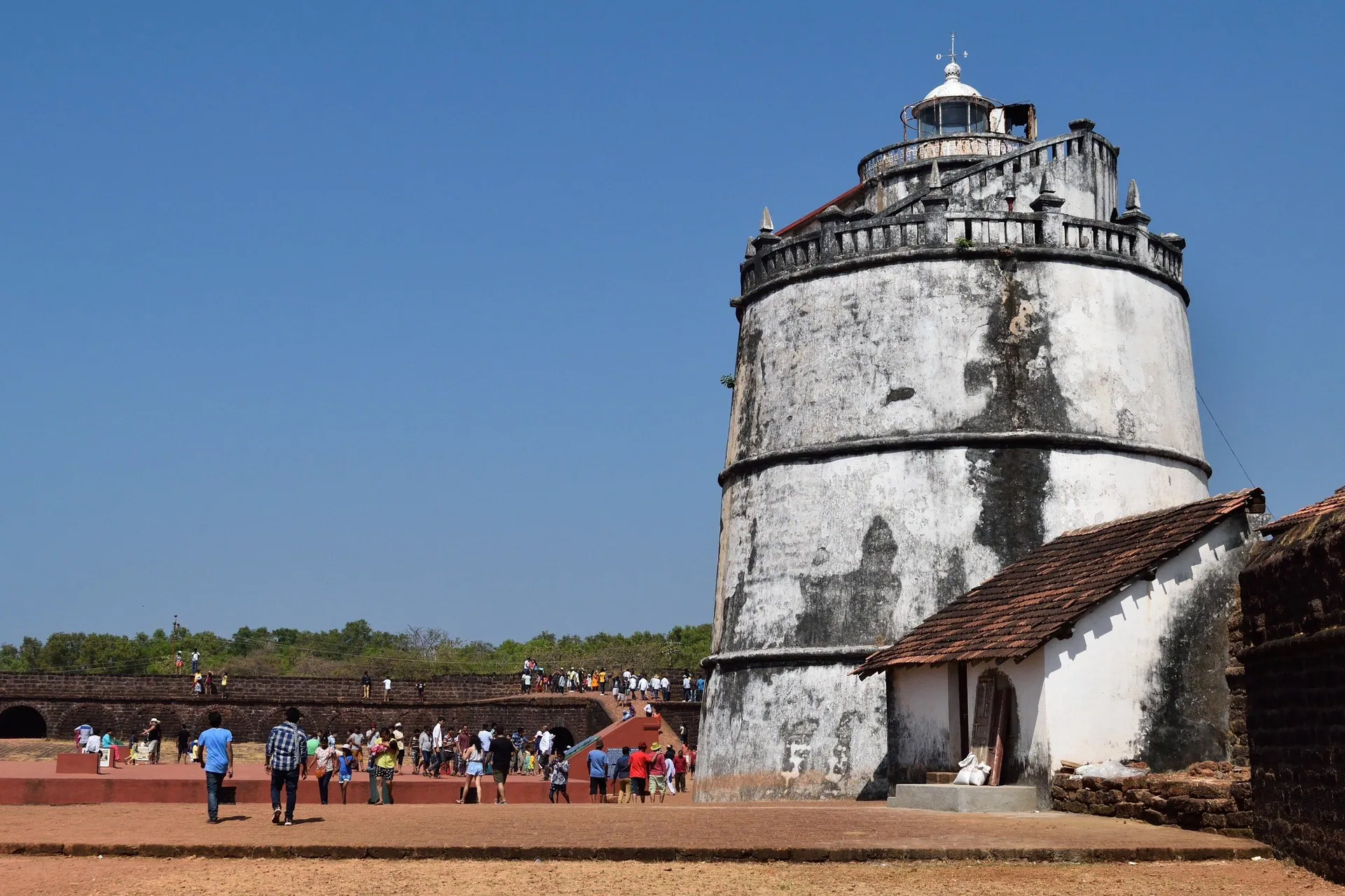 The weathered old lighthouse of Fort Aguada under a deep blue sky, visitors strolling below, Candolim, North Goa