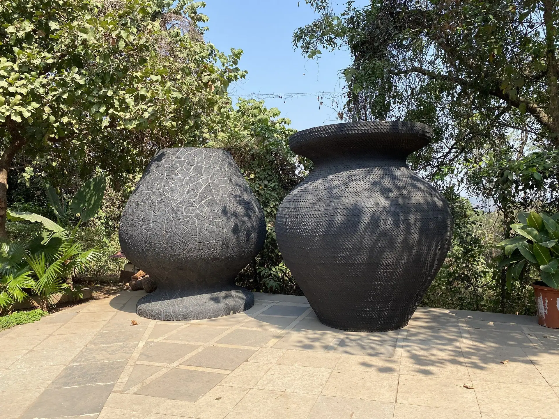 Two giant dark pot sculptures on a paved terrace in the tree-shaded garden of the Museum of Goa, Pilerne