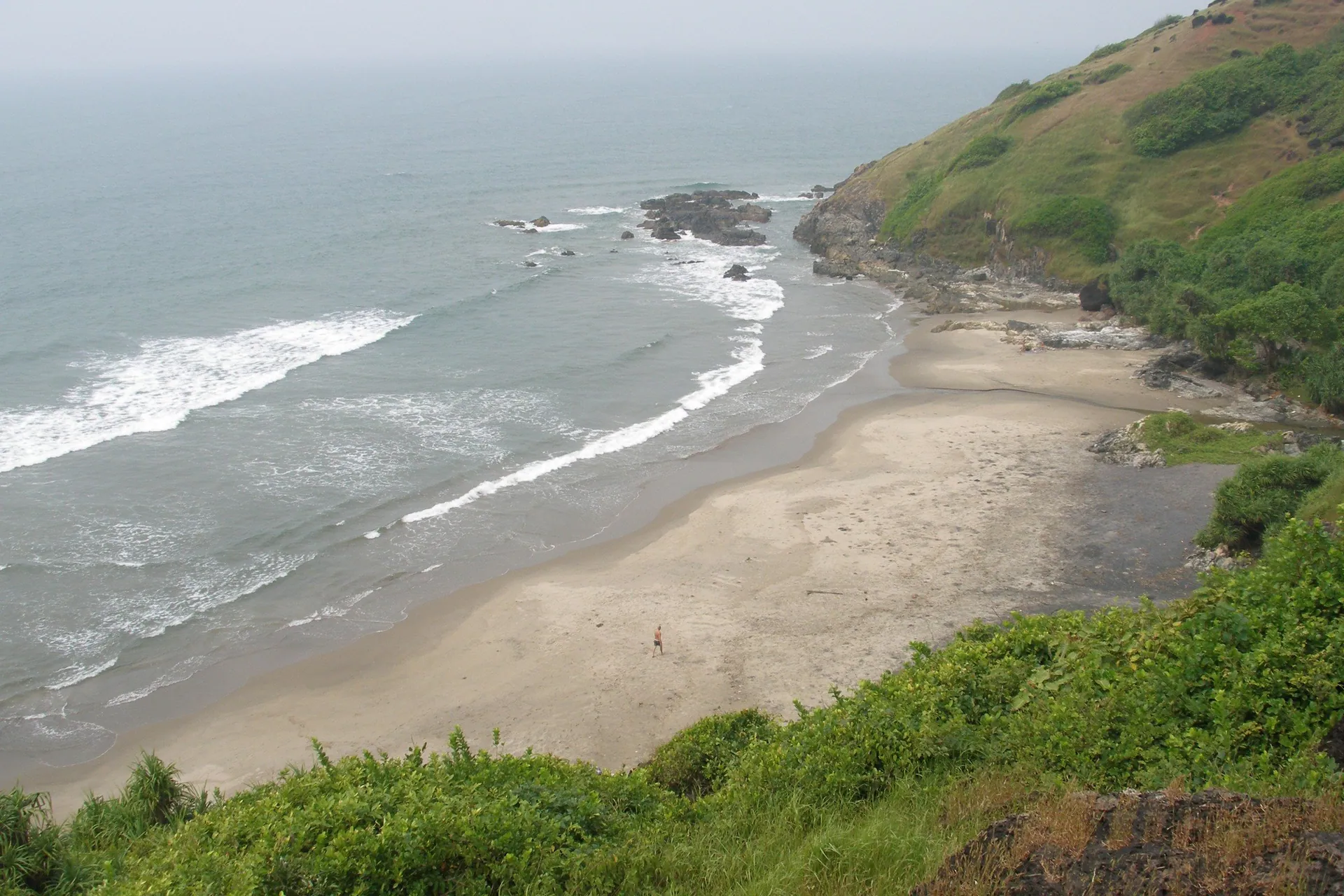 A quiet sandy cove at Vagator below grassy cliffs, with surf rolling in from the Arabian Sea